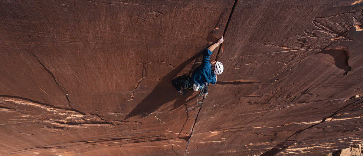 A high-angle action shot of a rock climber scaling a massive red sandstone cliff face, wearing a blue jacket and white helmet.