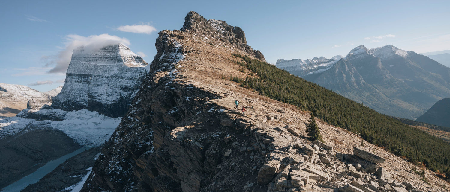 Two hikers made small by the scenic mountain landscape featuring a rugged rocky peak in the foreground, snow patches, distant mountains, and a clear blue sky.