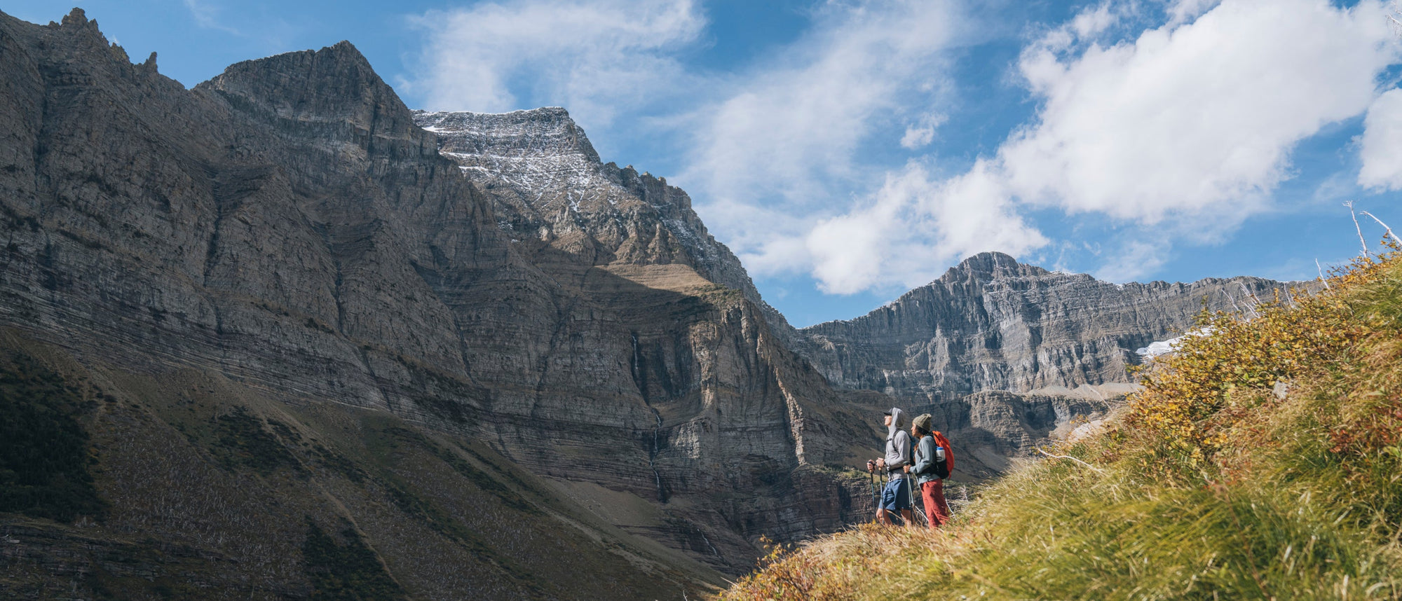A scenic mountain landscape with steep rocky peaks under a blue sky with white clouds, with two small hikers visible on a grassy slope in the foreground.