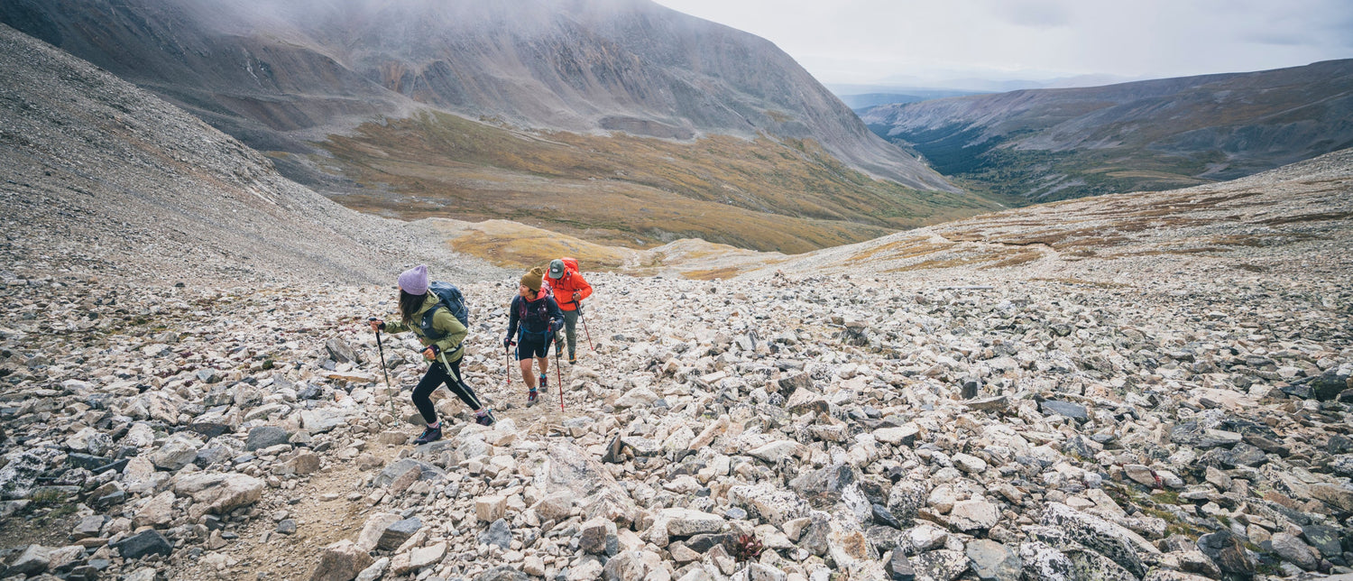 Three hikers with backpacks and trekking poles ascend a rocky mountain path in a vast, misty valley landscape.