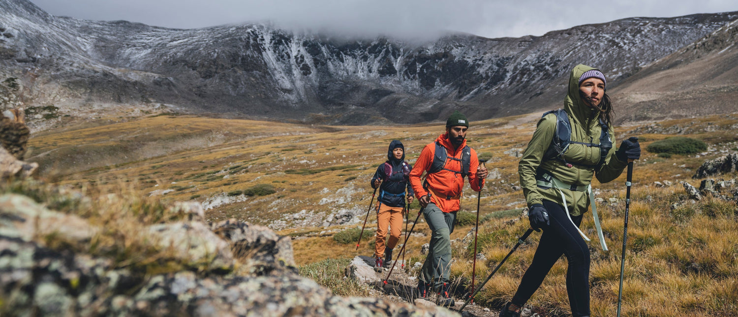Three hikers wearing Black Diamond gear trek through a rocky, grassy mountain landscape under a cloudy sky.