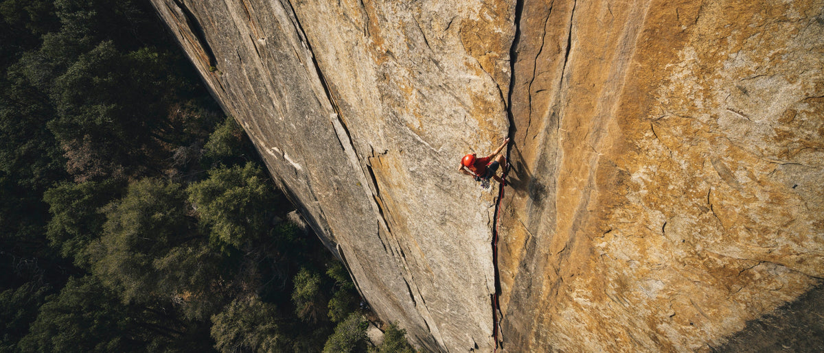 A high-angle photograph of a rock climber scaling a massive, sheer cliff face with a forest visible below.
