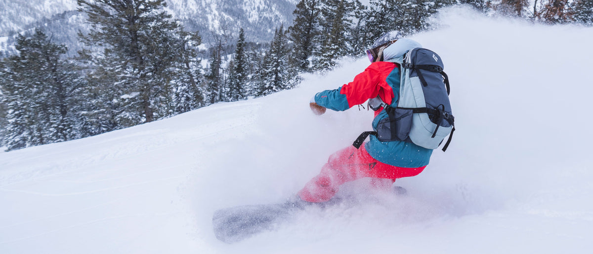 A snowboarder carving through deep powder snow on a mountain slope, wearing a bright red and teal snowsuit with a large gray backpack, with snowy trees and mountains in the background.