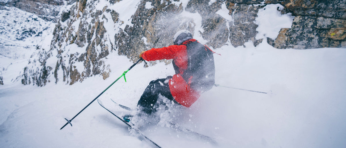 A skier in a red and black jacket carving through deep powder snow with rock walls in the background.