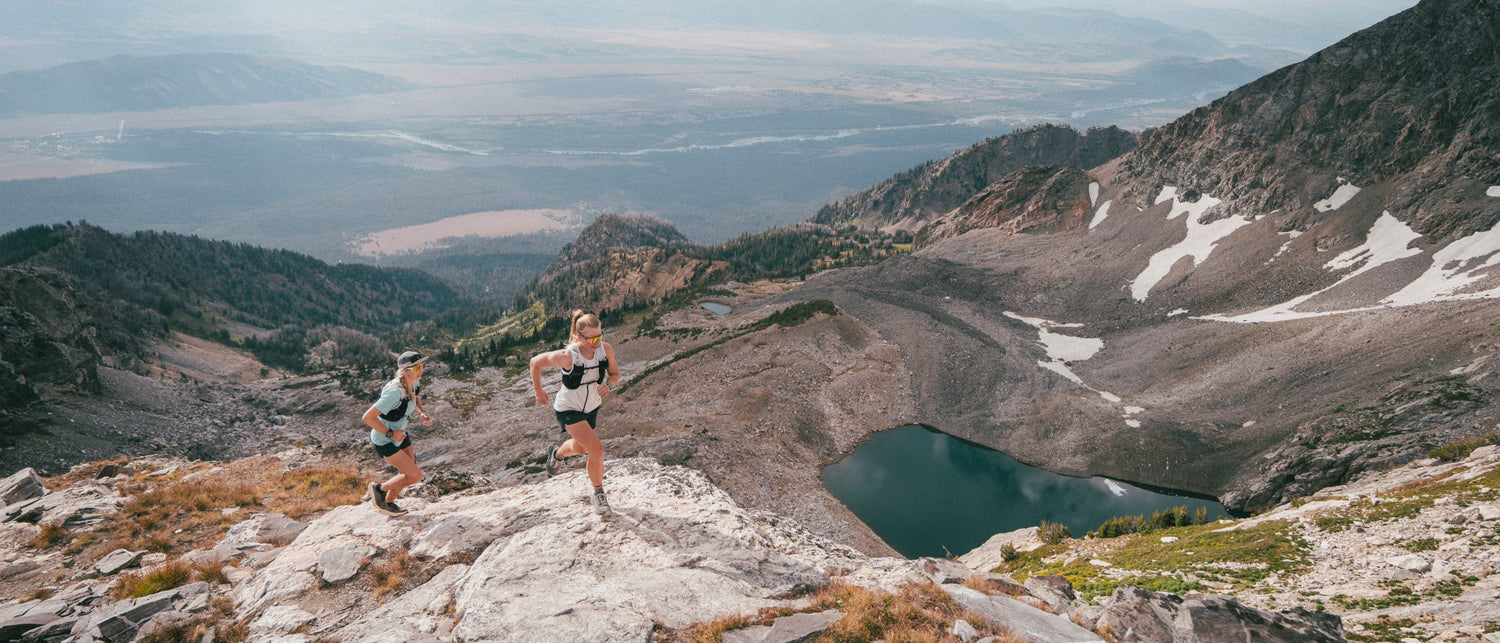 Two trail runners on a rocky mountain ridge overlooking a vast valley, with a small lake and patches of snow in the background.