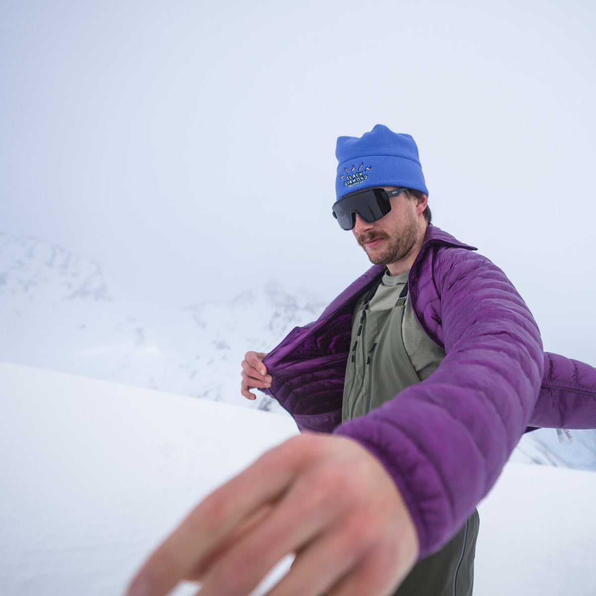 A skier pulls on a purple puffy jacket on the skin track. 