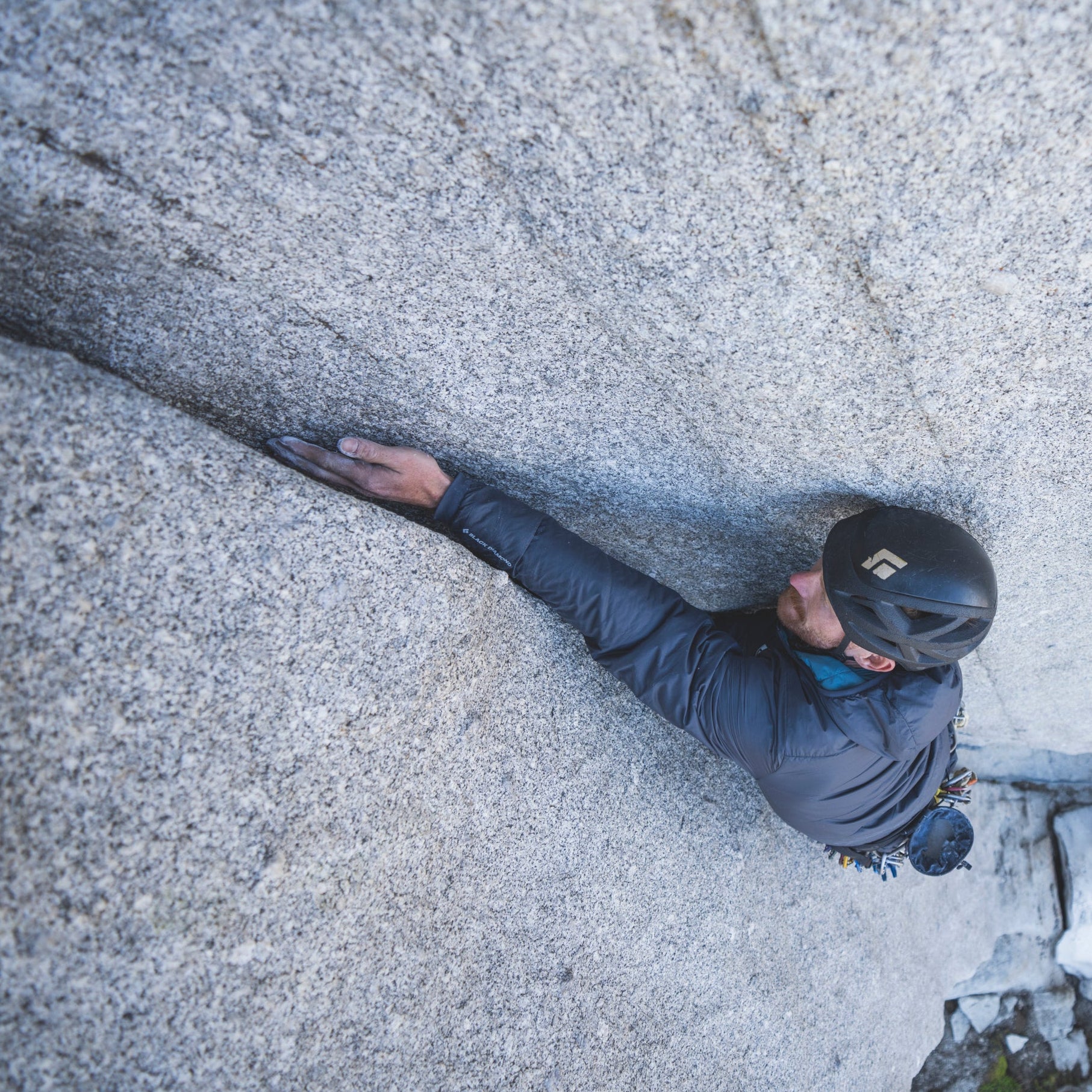 A high-angle shot of a rock climber wearing a black helmet and dark gray climbing jacket, scaling a massive textured gray granite wall.