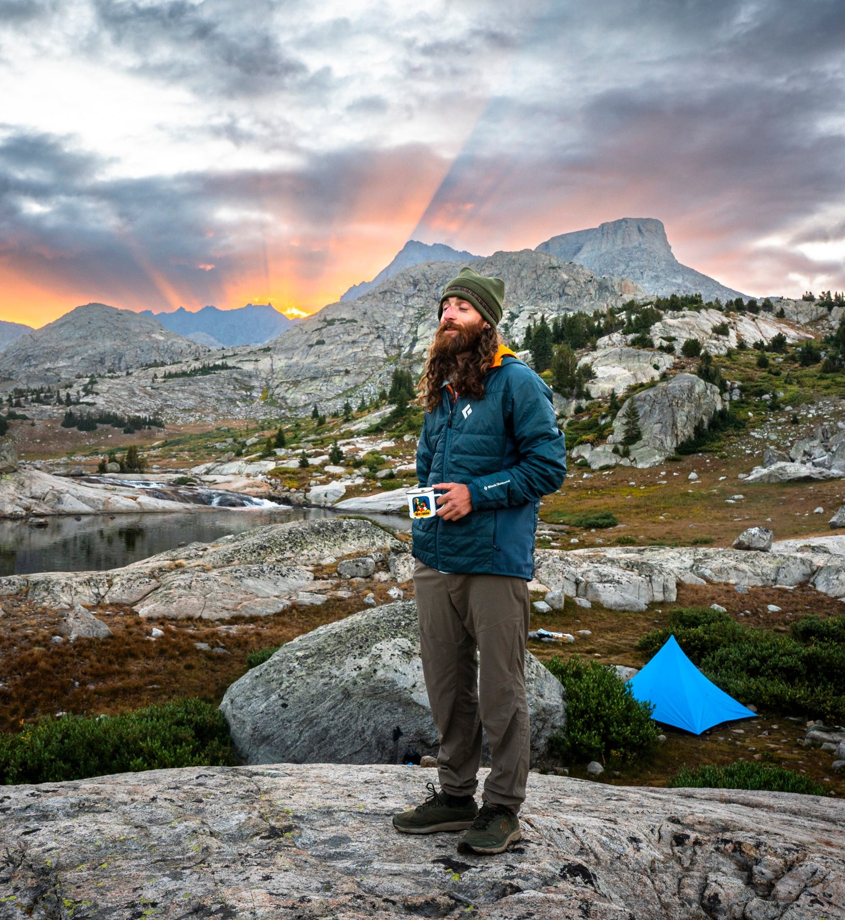 A person stands on a rocky mountain ledge holding a mug, with a mountain lake, rocky peaks, and a dramatic sunset sky in the background. A bright blue tent is pitched nearby on the ground.