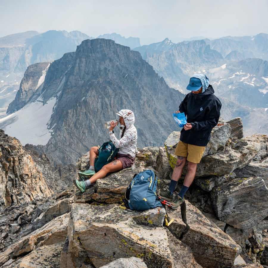 Two hikers on top of a summit having a snack, a mountain vista behind them.